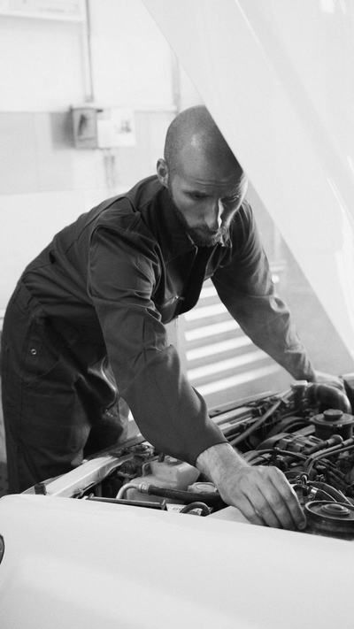 Mechanic in coveralls leaning into an engine bay under a raised hood
